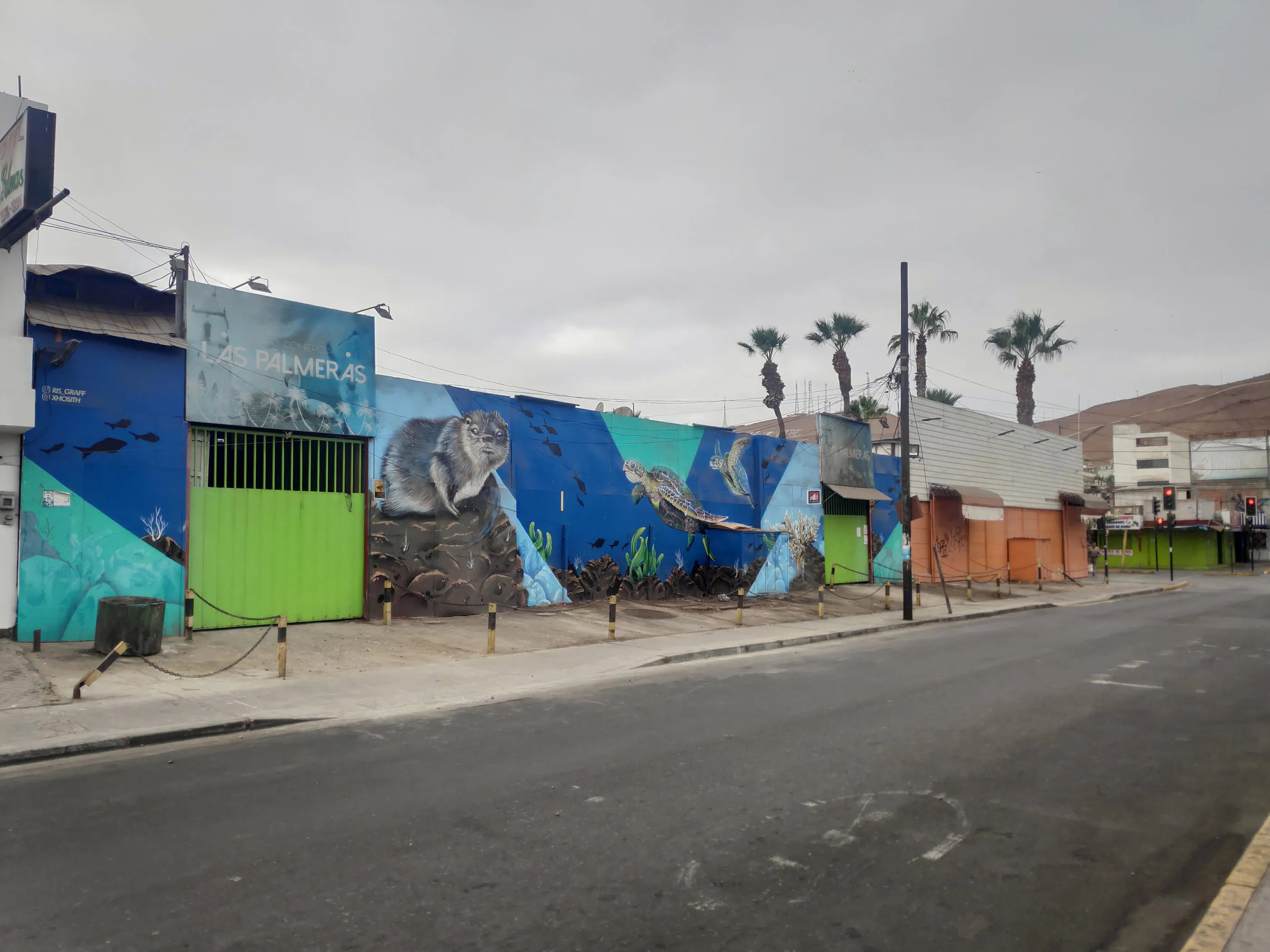 A long, colourful street mural beside a palm-lined harbour road